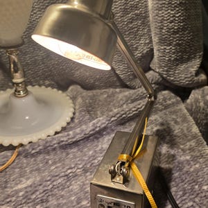 May include: A silver-toned desk lamp with an adjustable arm and a rectangular base. The lamp has a classic design with a pivoting head and a visible lightbulb. A yellow ribbon is tied around the base. The lamp is on a gray surface.