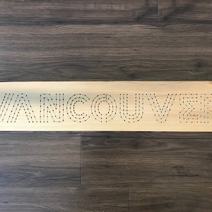 May include: Wooden sign with the word "VANCOUVER" spelled out in black dots. The sign is rectangular and made of light-colored wood. The background is a dark wood grain.
