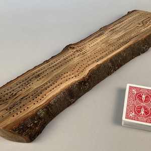 Può includere: Un tabellone di cribbage in legno con bordo naturale, con fori praticati per pioli e un mazzo di carte da gioco. Il tabellone è realizzato in legno di colore chiaro con motivi a venature scure. Sono inseriti pioli neri, rame e argento.