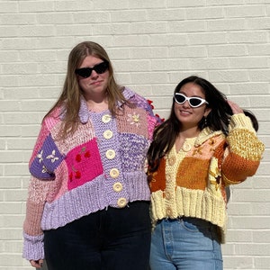 May include: Two people wearing colorful patchwork cardigans. The cardigan on the left is purple, pink, and white with embroidered flowers and cherries. The cardigan on the right is yellow, orange, and brown with embroidered flowers. Both cardigans are cropped and have button closures.