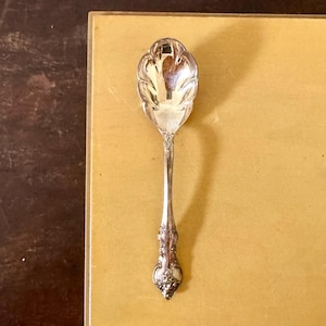 May include: A silver spoon with an ornate handle and a decorative, scalloped bowl. The spoon is placed on a yellow surface, with a dark brown surface and white-paneled wall in the background.