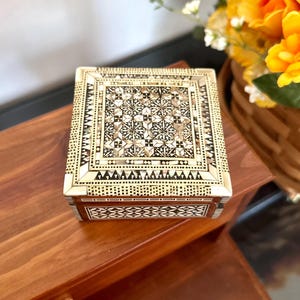 May include: A square wooden box featuring intricate geometric inlay work on the lid and sides. The lid displays a central pattern of star-like shapes surrounded by borders of small squares and triangles in cream, brown, and dark wood tones.