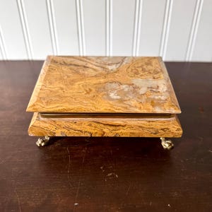 May include: A small, square jewelry box with a marbled, tan and brown stone exterior. The box sits on four small, gold-colored feet. The lid is flat and the box appears to be made of stone or a similar material.
