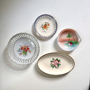 May include: Four decorative vintage plates with floral designs. Includes a round plate with a blue border and floral center, a small handled dish with a landscape scene, a pierced plate, and an oval plate with a pink flower.