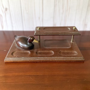 May include: A vintage wooden pipe stand with a duck figurine. The stand is dark brown with a rectangular base and three circular cutouts. The duck is also dark brown with a yellow beak. A small shelf is attached to the stand.