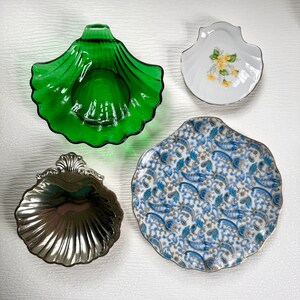 May include: Four decorative shell-shaped dishes. A vibrant green glass dish, a silver-colored metal dish, and two ceramic dishes. One ceramic dish has a white background with yellow floral accents, and the other has a blue and white paisley pattern.
