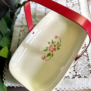 May include: Cream-colored rectangular serving dish with a red handle. The dish features a floral design in pink and green. The handle is made of red metal. The dish is sitting on a white lace tablecloth.
