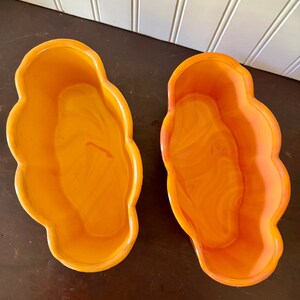 May include: Two orange, cloud-shaped planters. The planters have a glossy finish and a marbled appearance. The planters are empty and sit on a dark brown surface. The background is a white wall with vertical paneling.