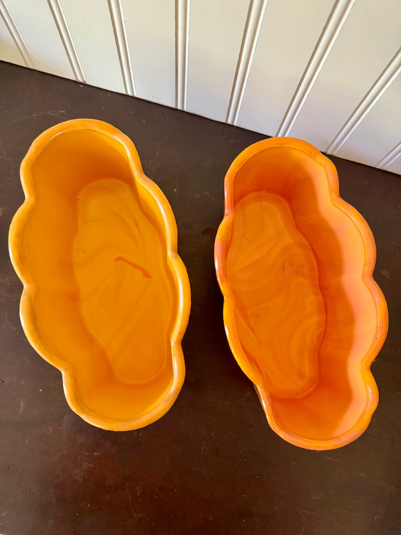May include: Two orange, cloud-shaped planters. The planters have a glossy finish and a marbled appearance. The planters are empty and sit on a dark brown surface. The background is a white wall with vertical paneling.
