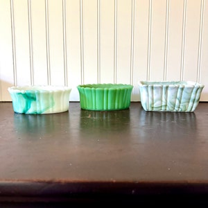 May include: Three small, rectangular glass dishes in shades of green and white. The dishes have a scalloped edge design. The dish on the left has a marbled green and white pattern. The middle dish is solid green. The dish on the right has a marbled green and white pattern.