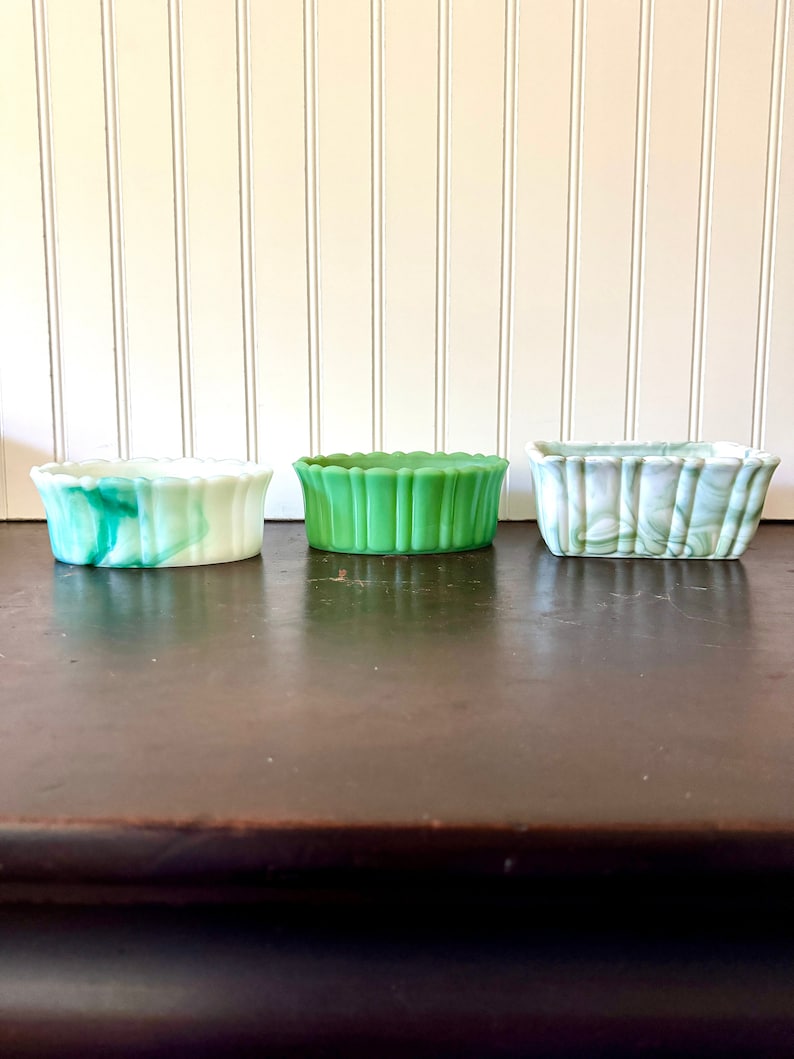 May include: Three small, rectangular glass dishes in shades of green and white. The dishes have a scalloped edge design. The dish on the left has a marbled green and white pattern. The middle dish is solid green. The dish on the right has a marbled green and white pattern.