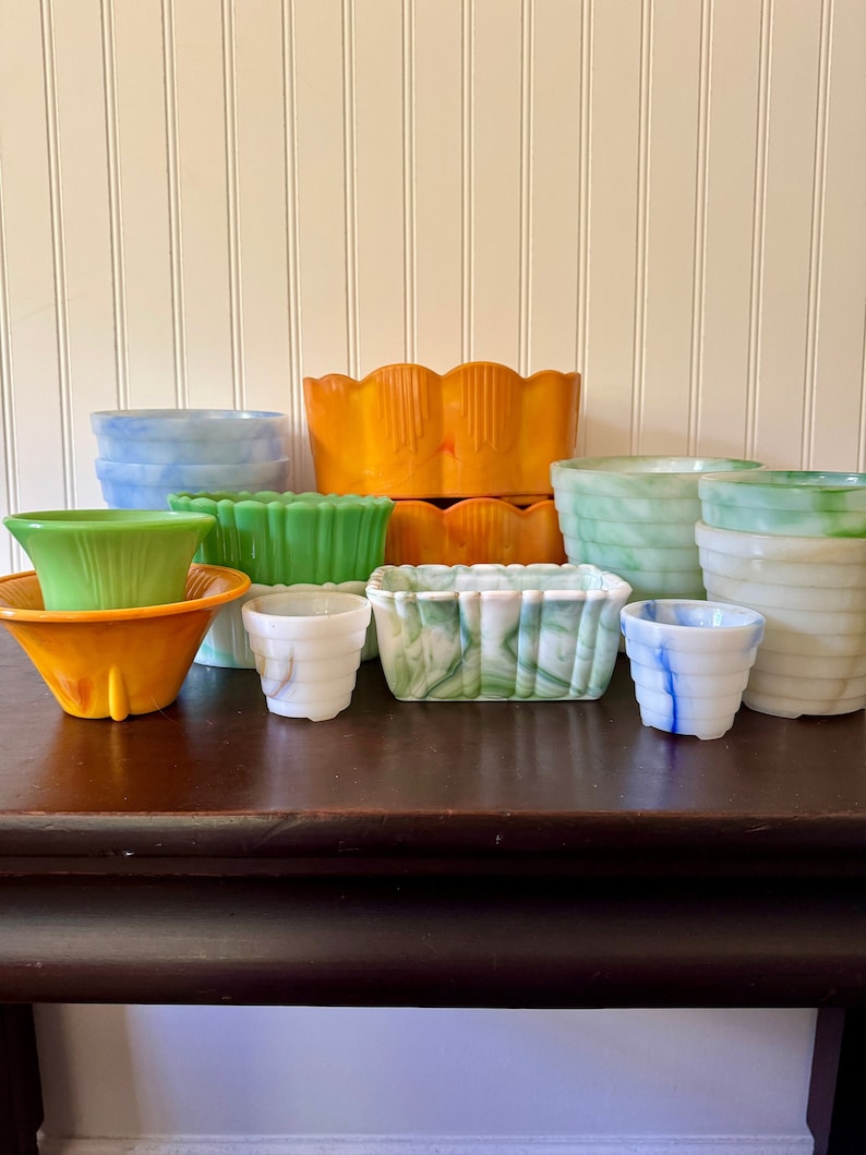 May include: A collection of colorful vintage planters in various shapes and sizes. The planters are in shades of blue, green, orange, and white. Some have ribbed or scalloped designs. The planters are arranged on a dark brown table against a white wall.