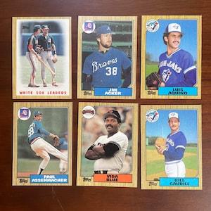 May include: Six vintage baseball cards featuring players from various teams. The cards have a gold border and include images of players like Jim Acker, Vida Blue, and Luis Aquino. The text on the cards includes team names and player names.