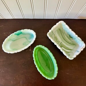 May include: Three decorative glass trays in shades of green and white. Two are oval-shaped with scalloped edges, one with a marbled effect. The third is rectangular with a scalloped edge and a marbled design. All are on a dark brown surface.