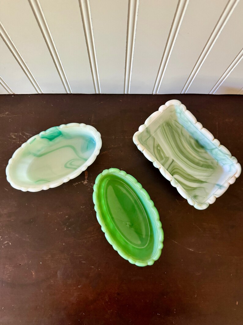 May include: Three decorative glass trays in shades of green and white. Two are oval-shaped with scalloped edges, one with a marbled effect. The third is rectangular with a scalloped edge and a marbled design. All are on a dark brown surface.
