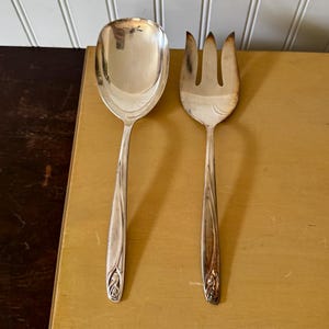 May include: A silver serving spoon and fork set. The spoon has a wide, oval bowl, and the fork has three tines. Both utensils have long, slender handles with a decorative floral design at the base.