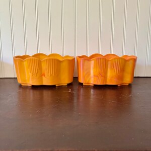 May include: Two orange rectangular planters with scalloped edges. The planters have a vertical design on the front and are sitting on a dark brown surface. The background is a white wall with vertical wooden panels.