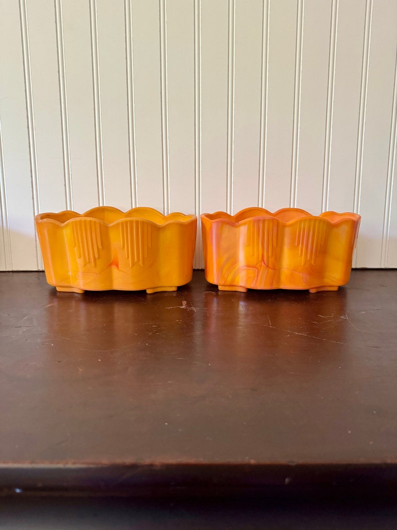 May include: Two orange rectangular planters with scalloped edges. The planters have a vertical design on the front and are sitting on a dark brown surface. The background is a white wall with vertical wooden panels.