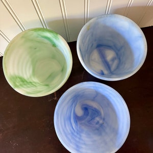 May include: Three decorative bowls with a marbled design. One bowl is green and white, another is light blue and white, and the third is a darker blue and white. The bowls are round and have a smooth, glossy finish. They are arranged on a dark brown surface.
