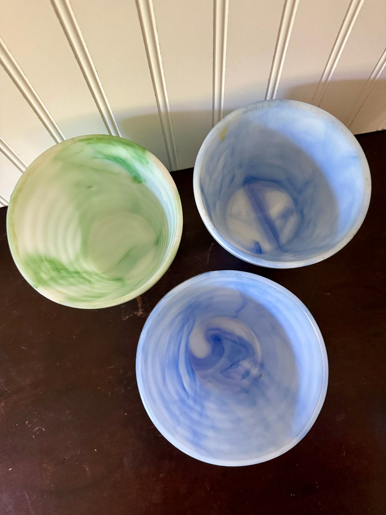 May include: Three decorative bowls with a marbled design. One bowl is green and white, another is light blue and white, and the third is a darker blue and white. The bowls are round and have a smooth, glossy finish. They are arranged on a dark brown surface.