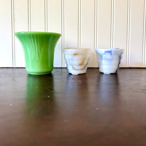 May include: Three decorative glass vases. A tall, flared green vase stands next to two smaller, cylindrical vases. One small vase is white with brown marbling, and the other is white with blue marbling. The vases are arranged on a dark brown surface.
