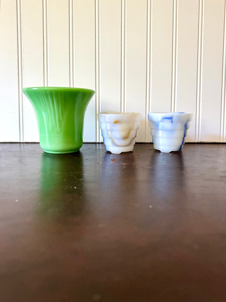 May include: Three decorative glass vases. A tall, flared green vase stands next to two smaller, cylindrical vases. One small vase is white with brown marbling, and the other is white with blue marbling. The vases are arranged on a dark brown surface.