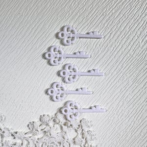 May include: Four white decorative keys with ornate floral designs are arranged on a textured white surface. A white lace doily is in the lower left corner. The keys have the word "LOVE" printed on them.