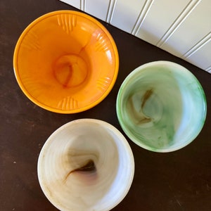 May include: Three vintage glass bowls in shades of orange, green, and white. The orange bowl has a textured rim. The bowls have a swirled pattern. The bowls are on a dark brown surface.