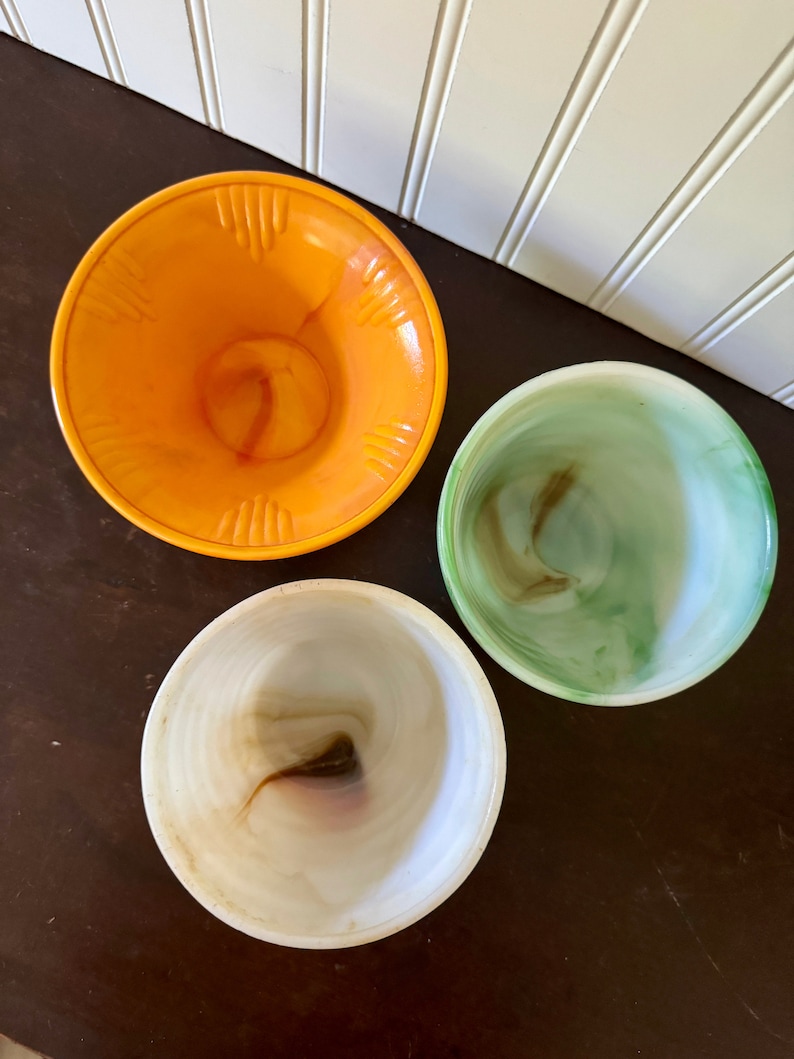 May include: Three vintage glass bowls in shades of orange, green, and white. The orange bowl has a textured rim. The bowls have a swirled pattern. The bowls are on a dark brown surface.
