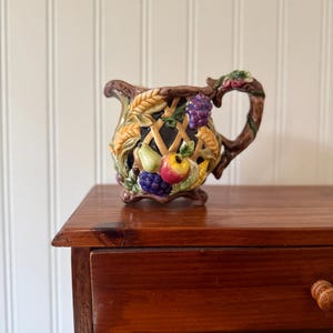May include: A decorative ceramic creamer with a basket weave design, featuring colorful fruit and wheat accents. The handle is shaped like a branch. The creamer is set on a wooden surface.