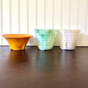 May include: Three decorative planters in a row. The first is orange, flared, and has a ridged texture. The second is green and blue, with a marbled effect. The third is white with brown marbling and a ridged texture. All are made of a translucent material.
