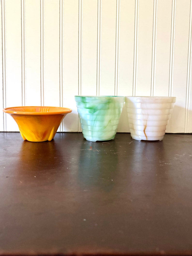 May include: Three decorative planters in a row. The first is orange, flared, and has a ridged texture. The second is green and blue, with a marbled effect. The third is white with brown marbling and a ridged texture. All are made of a translucent material.