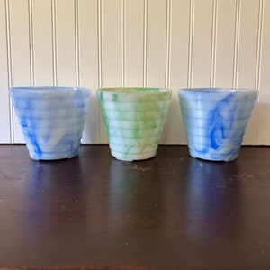 May include: Three decorative, tiered planters in shades of blue, green, and white. The planters have a marbled appearance and are arranged on a dark brown surface. The background is a white wall with vertical paneling.
