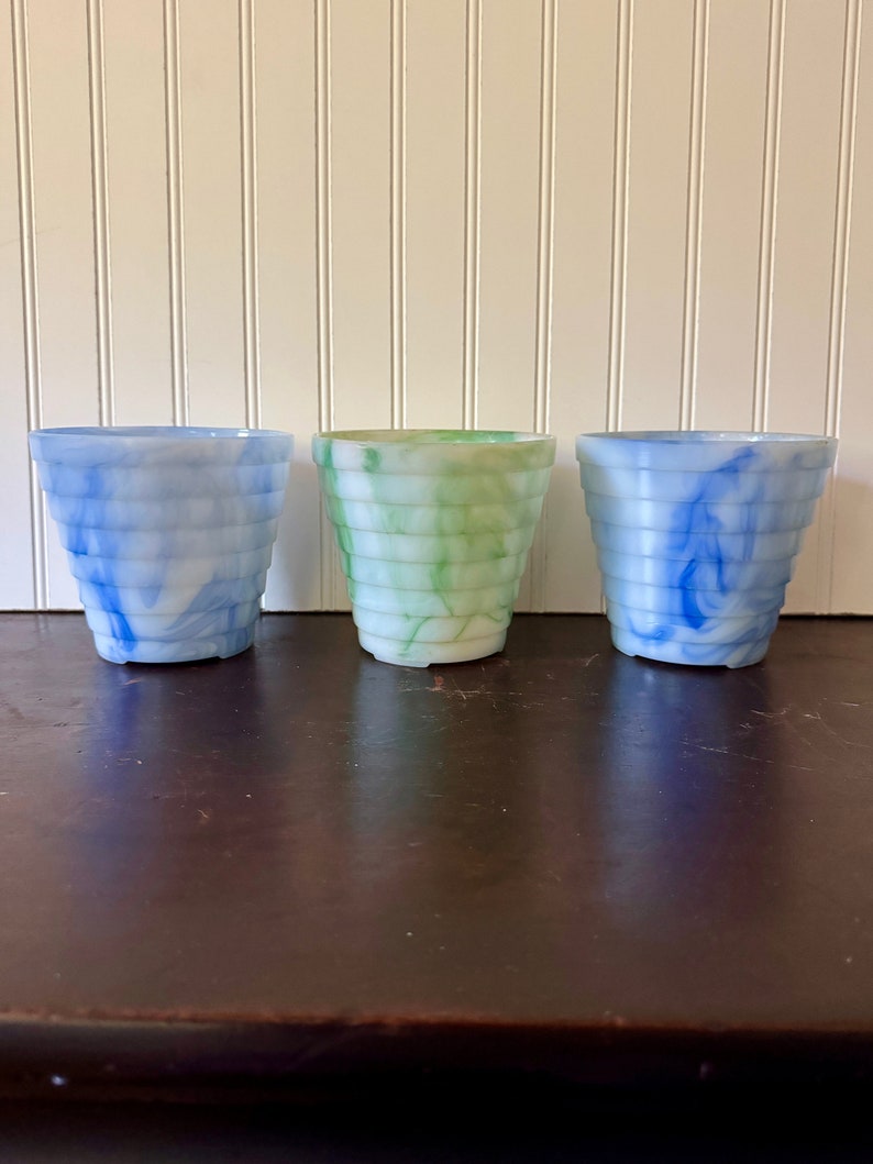 May include: Three decorative, tiered planters in shades of blue, green, and white. The planters have a marbled appearance and are arranged on a dark brown surface. The background is a white wall with vertical paneling.