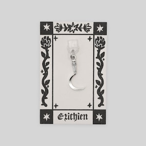 May include: A silver crescent moon charm earring on a decorative card. The card has a black and white floral border and the word "Ezithien" printed at the bottom. The earring is attached to a clear plastic backing.