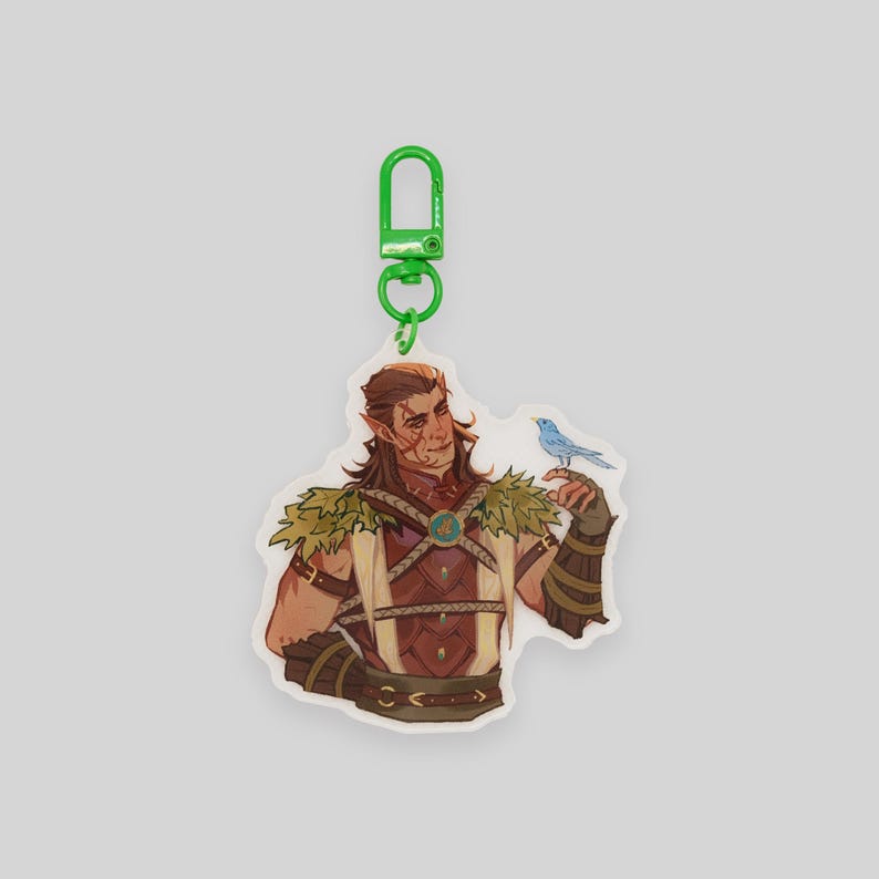 May include: A white acrylic keychain featuring a cartoon illustration of a person with brown hair and a blue bird. The person is wearing a brown leather vest with straps and a green leafy collar. The keychain has a green metal ring.