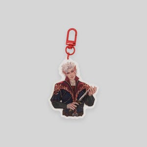 May include: A white acrylic keychain featuring a character with white hair, wearing a dark blue and red outfit with gold accents, holding a silver dagger. The keychain has a red metal clasp.