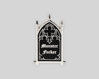May include: A silver-toned metal enamel pin shaped like a gothic arched window. The pin features a black background with white decorative tracery at the top and the text "Monster Fucker" written in a white gothic-style font in the center.