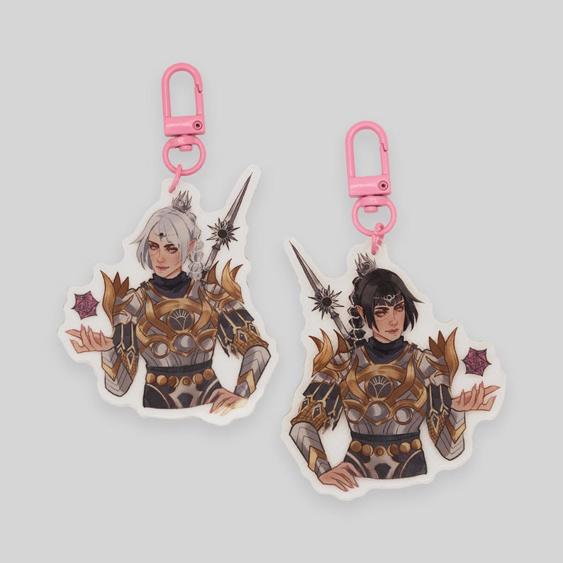 May include: Two white acrylic keychains with pink metal clasps. Each keychain features a different illustration of a woman in armor holding a glowing orb. The woman on the left has white hair and the woman on the right has black hair.