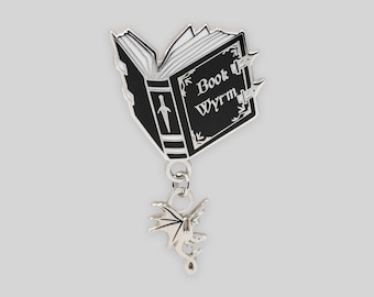 May include: A black and silver metal enamel pin shaped like an open book with the text Book Wyrm on the cover. The book features a white sword icon on the spine and a small silver dragon charm dangling from the bottom by a metal jump ring.