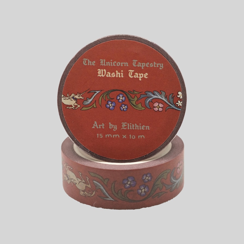May include: A roll of terracotta-colored washi tape featuring a repeating pattern of a unicorn and floral vines. The label reads The Unicorn Tapestry Washi Tape, Art by Elithien, 15 mm x 10 m. The tape has a matte finish with detailed illustrations.