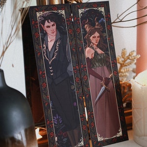 May include: Two bookmarks with a black background and gold trim. The bookmarks feature illustrations of a man and a woman in elaborate clothing. The man is wearing a black suit with gold embroidery and the woman is wearing a long, flowing dress with a green bodice. Both bookmarks have a red berry pattern around the border.