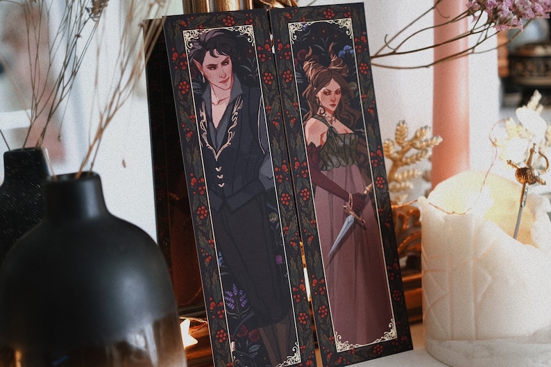 May include: Two bookmarks with a black background and gold trim. The bookmarks feature illustrations of a man and a woman in elaborate clothing. The man is wearing a black suit with gold embroidery and the woman is wearing a long, flowing dress with a green bodice. Both bookmarks have a red berry pattern around the border.