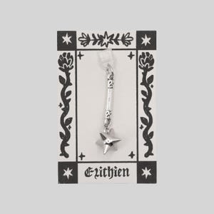 May include: A silver-toned metal charm featuring a long, textured cylindrical stem topped with a five-pointed star. The charm is attached to a white rectangular display card decorated with black floral borders and the text Elithien at the bottom.