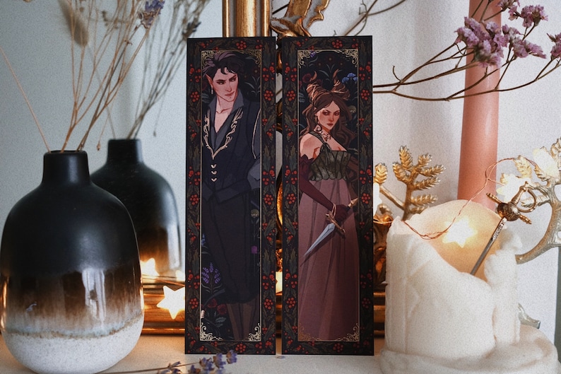 May include: Two bookmarks with intricate designs featuring a man and a woman in elaborate clothing. The bookmarks are set against a dark background with floral patterns and red berries. The man is wearing a black suit with gold accents and the woman is wearing a long, flowing dress with a dagger.