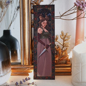 May include: A bookmark featuring a woman in a long, burgundy dress holding a dagger. The background is a dark, floral pattern with red berries.