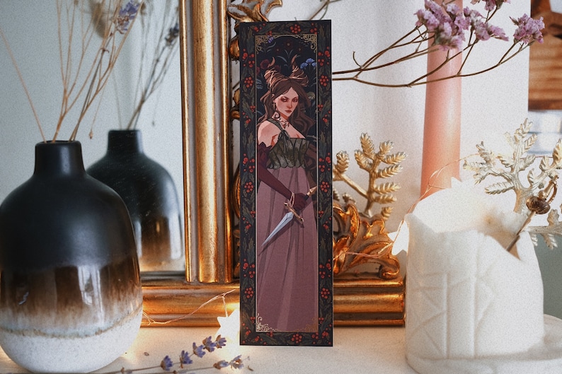 May include: A bookmark featuring a woman in a long, burgundy dress holding a dagger. The background is a dark, floral pattern with red berries.