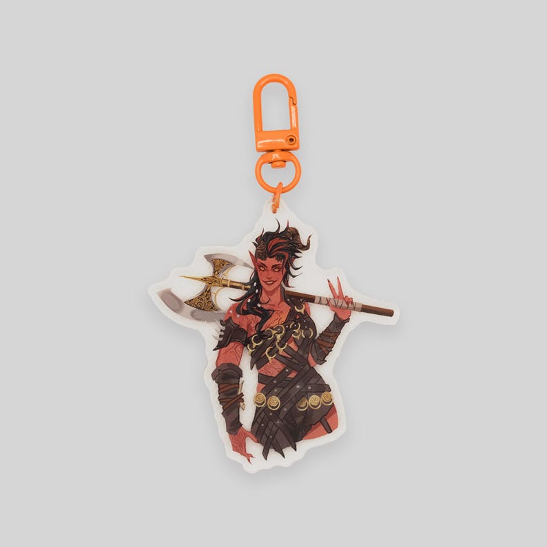 May include: A keychain featuring a colorful illustration of a warrior with red skin, horns, and a large axe. The keychain is made of clear acrylic and has an orange metal clasp.