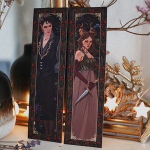 May include: Two bookmarks featuring illustrations of a man and a woman in elaborate clothing. The man is wearing a black suit with gold accents and the woman is wearing a long, flowing dress with a dagger. Both bookmarks have a dark background with a gold border and red berries.
