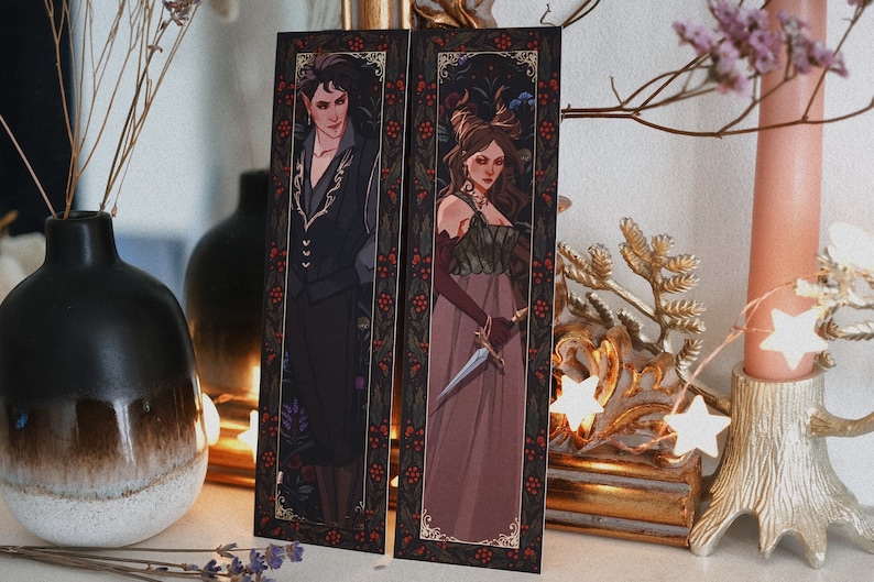 May include: Two bookmarks featuring illustrations of a man and a woman in elaborate clothing. The man is wearing a black suit with gold accents and the woman is wearing a long, flowing dress with a dagger. Both bookmarks have a dark background with a gold border and red berries.
